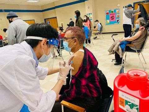 A masked clinician administering a vaccine to a seated community member.