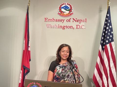 Prabha Bhattarai speaking at a podium between the Nepali and American flags at the Embassy of Nepal.