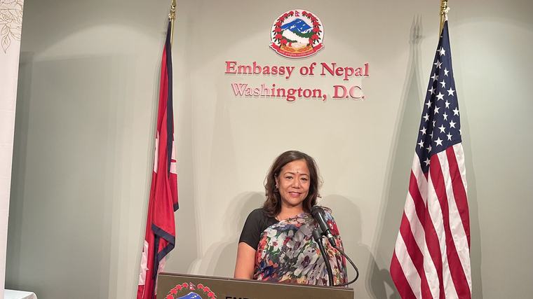 Prabha Bhattarai speaking at a podium between the Nepali and American flags at the Embassy of Nepal.