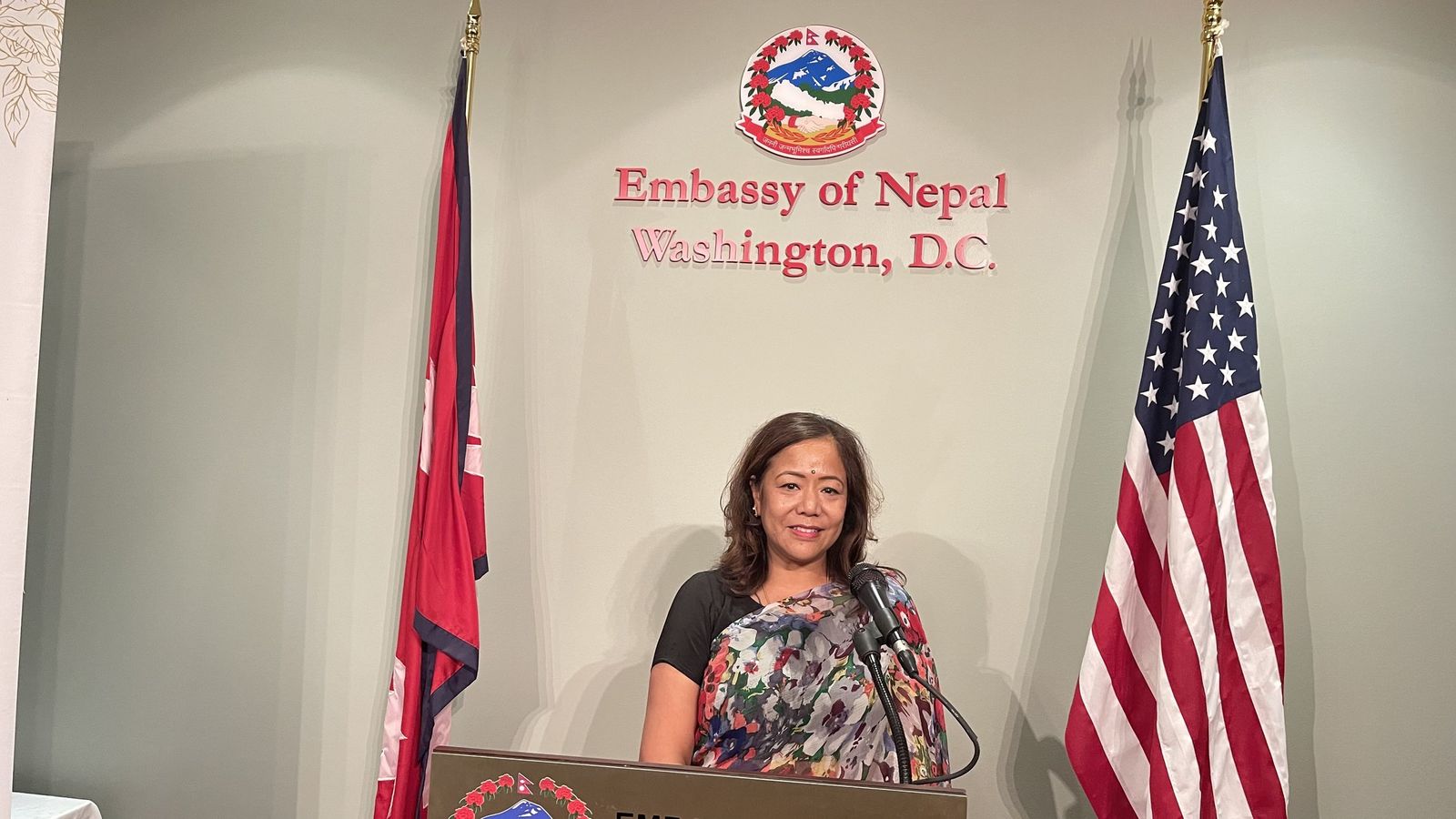 Prabha Bhattarai speaking at a podium between the Nepali and American flags at the Embassy of Nepal.